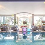 A room with a swimming pool in the foreground, large windows in the background, casual furniture and LED cube seating, floral decorations, large decorative pillars and a bar to one side. Set up for a milestone birthday celebration.